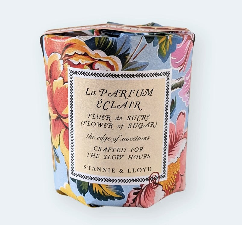 A square glass jar candle wrapped in a colorful floral paper sleeve, with a cream paper label in the center reading 'La PARFUM ÉCLAIR' and 'STANNIE & LLOYD' at the bottom, photographed against a plain white background.