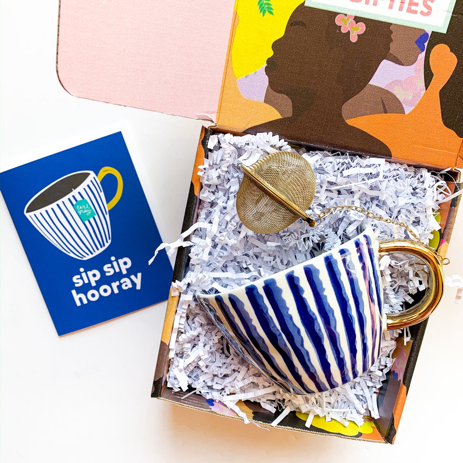 gift box filled with white crinkle paper featuring a striped ceramic tea mug with a gold sphere tea infuser and a greeting card that says Sip Sip Hooray